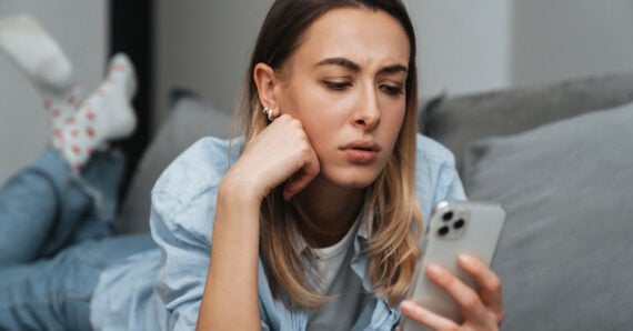 A woman lies on a couch, propped up on her elbow, looking at her smartphone with a concerned or puzzled expression. She is wearing a light blue shirt and jeans.