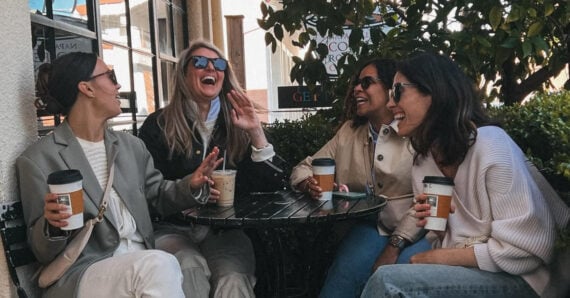 Four women sit together at an outdoor table, laughing and holding coffee cups, enjoying a casual conversation on a sunny day. Bushes and a building are in the background.