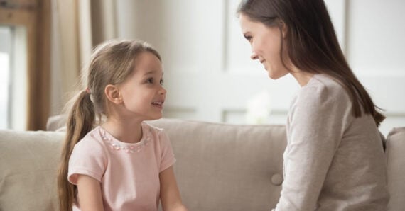 A young girl and a woman sit on a couch facing each other, smiling warmly and making eye contact, suggesting a friendly or caring conversation in a cozy, well-lit room.