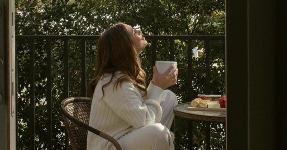 A person in white loungewear sits on a balcony, holding a mug and looking up, bathed in warm sunlight. Trees and greenery are visible behind them, and a small table with food is nearby.