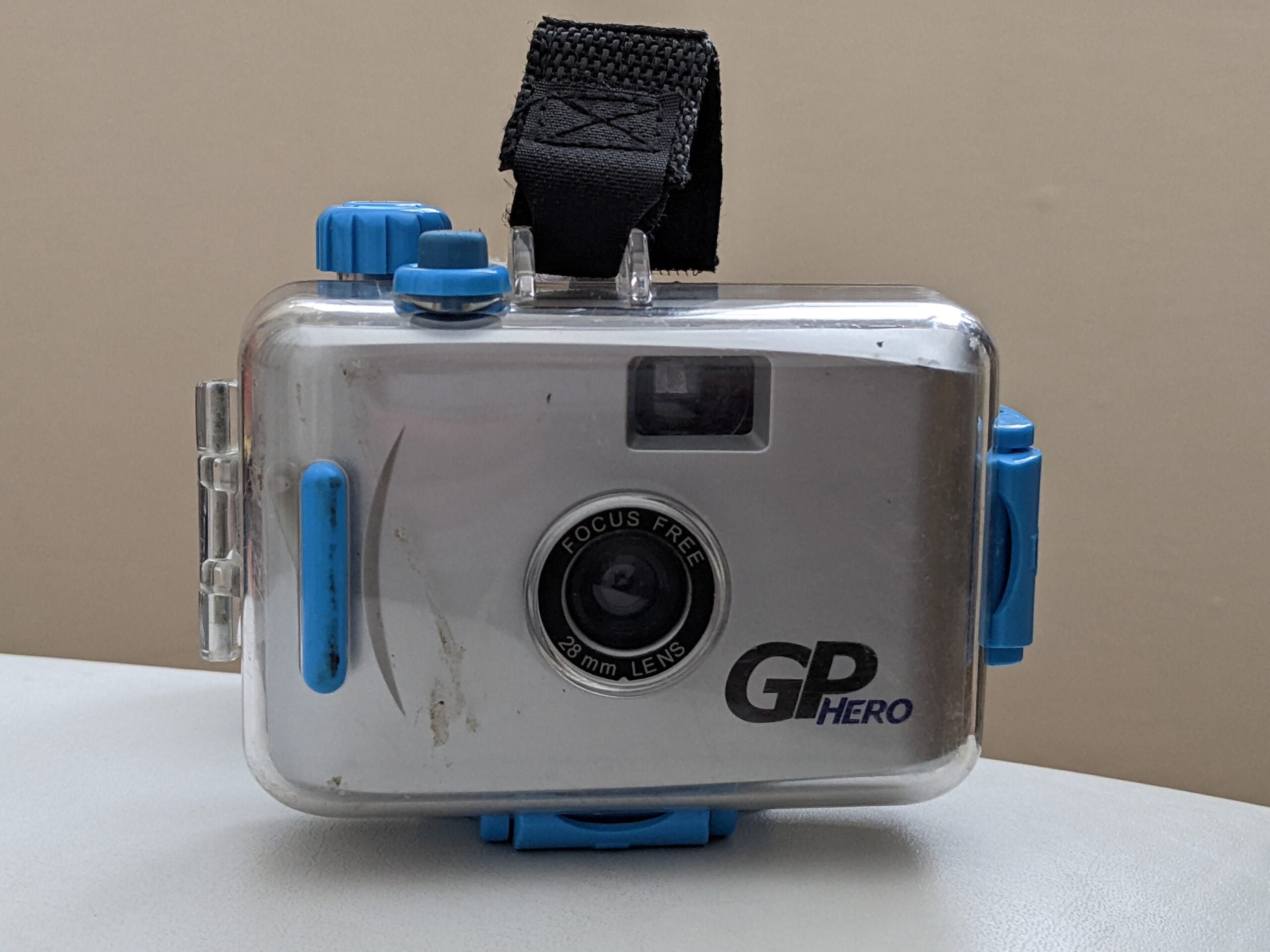 A silver GP Hero camera with a 28mm lens, blue controls, a black wrist strap, and a clear protective case, resting on a light surface against a plain beige background.