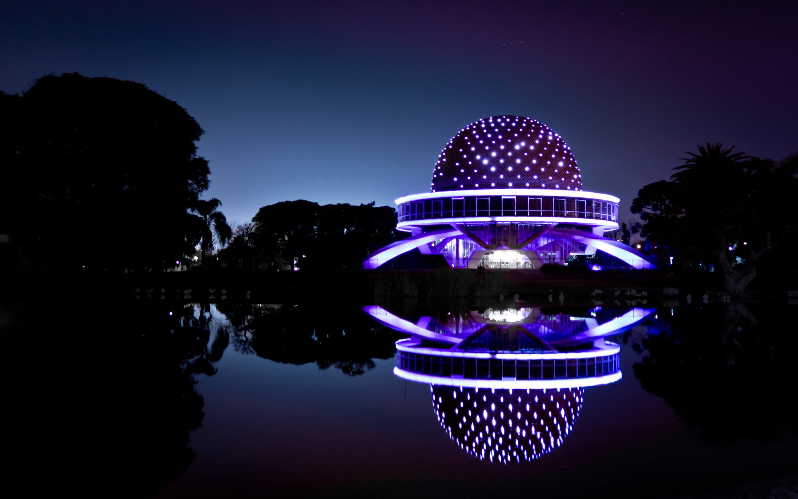 A futuristic, dome-shaped building illuminated with purple and white lights is reflected in a calm body of water at night, with silhouettes of trees in the background.
