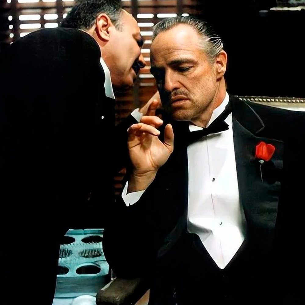 A man in a tuxedo sits pensively as another man leans in to whisper in his ear, set in a dim room with blinds casting shadows in the background. The seated man wears a red rose on his lapel.