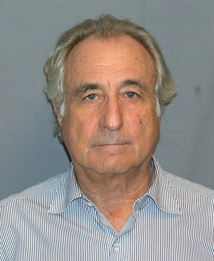 Headshot of an elderly man with gray-brown hair, wearing a blue and white striped collared shirt, photographed against a gray background in a style resembling an official mugshot.