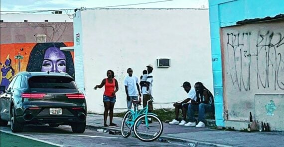 A group of people stand and sit on a sidewalk near a blue building, with a teal bicycle and a black car parked nearby. A colorful mural of a woman's face is painted on a wall in the background.