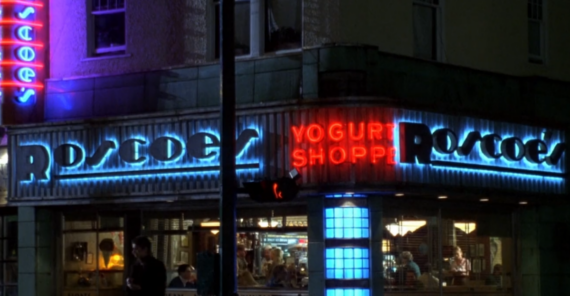 A night view of Roscoe's Yogurt Shoppe, with bright blue and red neon signs, large windows, and people sitting inside. The exterior is lit by neon lights, creating a vibrant urban atmosphere.
