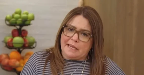 A woman with straight brown hair and glasses wears a striped shirt and layered necklaces, standing indoors with a fruit stand holding apples and oranges in the background.