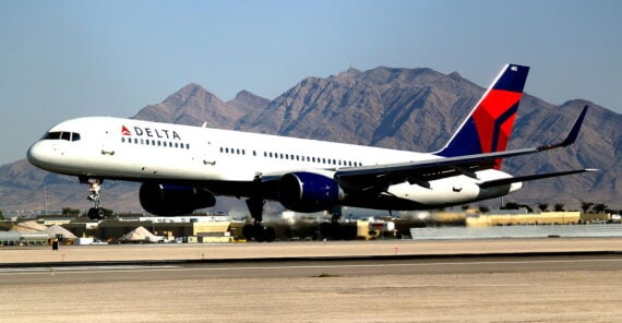 A Delta Air Lines airplane is landing on a runway with smoke rising from its tires. Mountains and airport buildings are visible in the background under a clear sky.