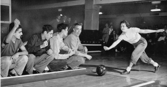 A young woman bowls while four young men sitting near the lane cheer her on in a vintage bowling alley. The group appears lively and engaged, with other people visible in the background.