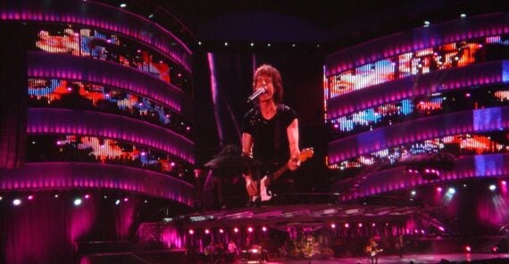 A musician plays guitar and sings on stage, with large, colorful LED screens displaying his image behind him. The stage is lit with vibrant pink and purple lights, creating a dynamic concert atmosphere.