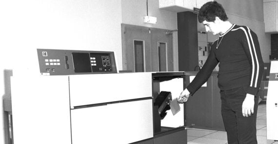 A person inserts a large magnetic disk into a vintage computer system in a retro, likely 1970s, data center setting. The room features tile floors and double doors in the background.