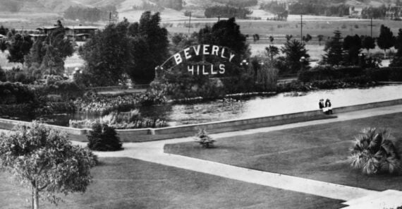 Historic black and white photo of the Beverly Hills sign beside a pond, surrounded by trees and landscaped gardens, with two people sitting on a bench near the water.