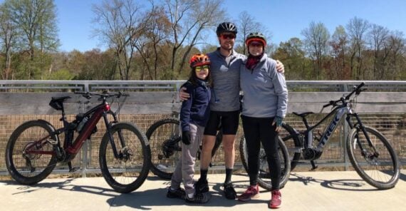 Three people wearing bike helmets stand smiling in front of a railing with two bicycles behind them, outdoors on a sunny day with trees in the background.