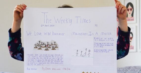A child holds up a handmade newspaper titled "The Weekly Times," featuring drawings of Warhammer figures, stick figures running a marathon, and handwritten articles and book review on decorated white paper.