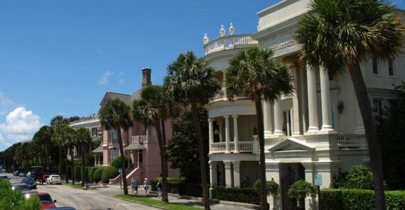 A street lined with historic mansions featuring large columns and balconies, palm trees along the sidewalk, and people walking on a sunny day.