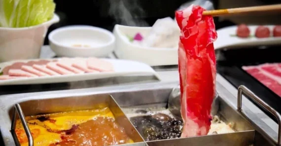 A close-up of a hot pot with four different broths, featuring a piece of raw beef being dipped with chopsticks. Plates of assorted meats and vegetables are arranged in the background. Steam rises from the boiling broth.