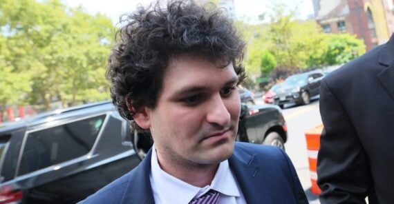 A man with curly brown hair in a suit and striped tie stands outdoors near parked cars and trees, looking slightly down and to the side.