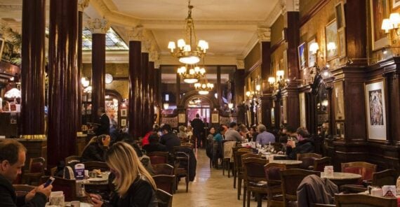 A lively, historic caf&eacute; with ornate chandeliers, dark wooden columns, and classic d&eacute;cor. People sit at tables, drinking and chatting, while warm lighting creates a cozy atmosphere. Paintings and mirrors adorn the walls.