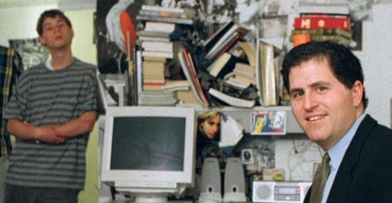Two men in a cluttered room with books, posters, a computer monitor, and speakers; one man in a suit smiles at the camera, while the other stands in the background wearing a striped shirt.