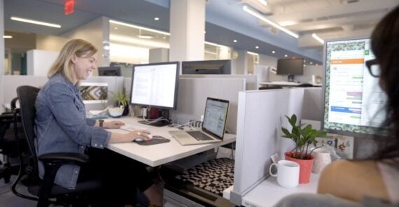 A woman sits at a desk in a modern open office, working on a computer with multiple monitors. There are plants, a mug, and office supplies on the desks, and other workstations in the background.
