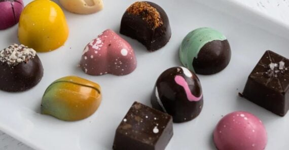 A white plate with assorted colorful, artisan chocolates in various shapes, including domes, squares, and ovals, each decorated with vibrant patterns and designs.