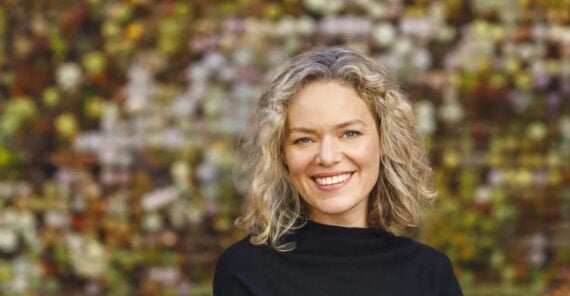 A woman with curly blonde hair and a black top smiles in front of a blurred background of flowers and greenery.