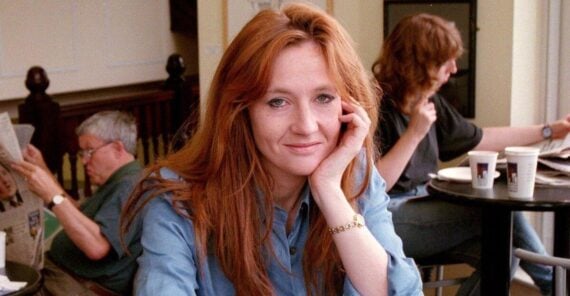 A woman with long red hair in a blue shirt sits at a table in a caf&eacute;, smiling at the camera. In the background, a man reads a newspaper and another woman sits at a table with two coffee cups.
