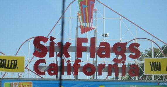 A young girl rides alone in a yellow roller coaster car on blue tracks, soaring through a theme park landscape with trees, greenery, and wooden support structures visible in the background.