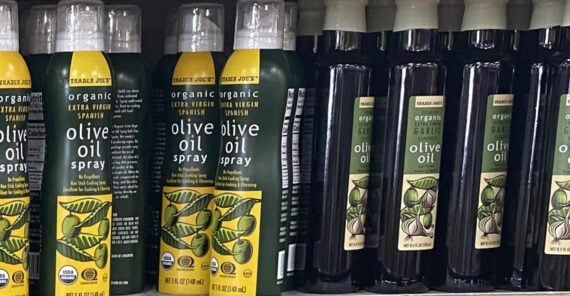 Bottles of Trader Joe&rsquo;s organic extra virgin olive oil spray are lined up on a store shelf next to bottles of Trader Joe&rsquo;s organic olive oil with a garlic label. The bottles feature green and yellow labels.