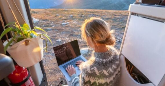A person with blond hair in a sweater sits in a van, working on a laptop, with the van door open to a sunny mountain landscape. A potted plant and fire extinguisher are visible inside the van.