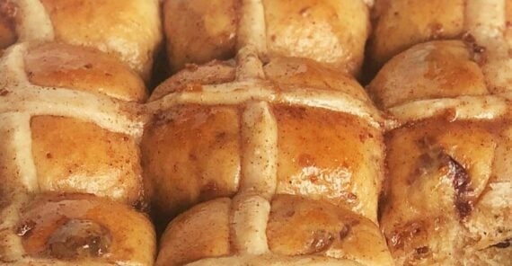 Close-up of freshly baked hot cross buns with a shiny, golden-brown glaze and white icing crosses on top. The buns are closely packed together and have visible pieces of dried fruit.