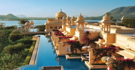 A luxurious palace-style hotel with domes and ornate architecture sits beside a long blue pool, lush gardens, and a lake, with mountains in the background under a clear sky.