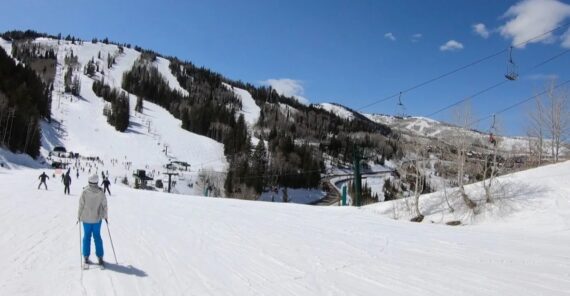 Skiers glide down a snowy mountain slope under a clear blue sky; ski lifts transport people up the hill, and pine-covered slopes surround the ski resort area.