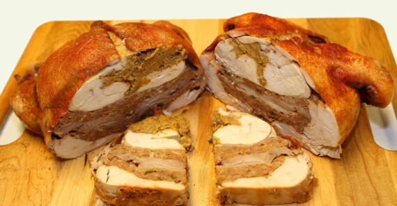 A sliced turducken on a wooden cutting board, showing layers of turkey, duck, chicken, and stuffing. Two thick slices are cut in front of the main portion, revealing the different meats and stuffing inside.