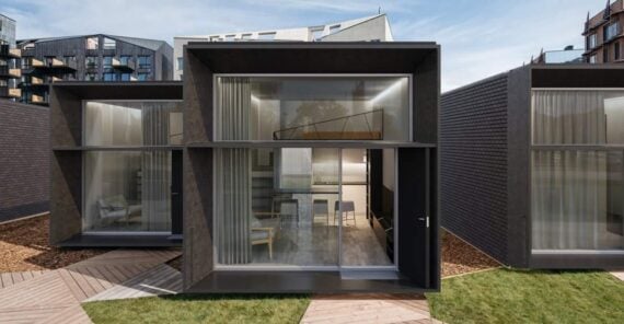 A modern, minimalist modular home with large glass windows, dark exterior panels, and visible interior furnishings, including a table and chairs, set among similar adjacent units with landscaped walkways and grass.