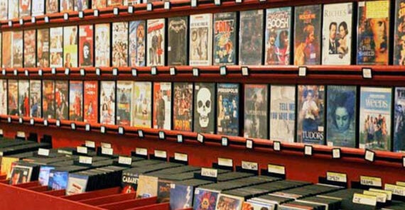 Rows of DVD and VHS cases are displayed on shelves in a video rental store, with movie titles visible on the covers. Black plastic cases are organized below for customer browsing.