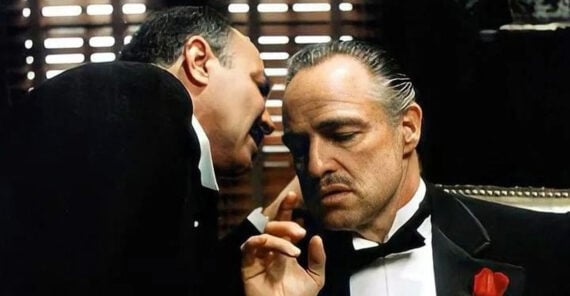 A man in a tuxedo whispers into the ear of another serious-looking man sitting in a dimly lit room, who wears a black suit, bow tie, and red rose on his lapel. Light filters through window blinds behind them.