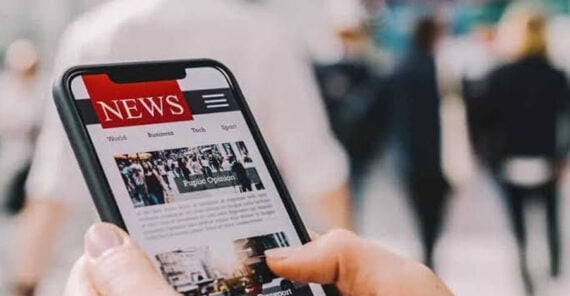 A person holds a smartphone displaying a news website, with sections like world, business, tech, and sports, while standing in a blurry, busy outdoor setting with people in the background.
