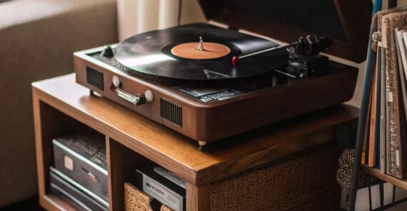A wooden record player with a vinyl record spinning sits on a wood shelf, surrounded by woven baskets, books, and albums in a cozy room setting.