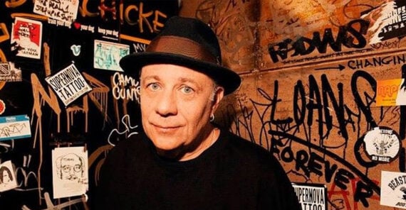 A man in a black shirt and hat stands in front of a graffiti-covered wall with various stickers, tags, and posters in a dimly lit urban setting.