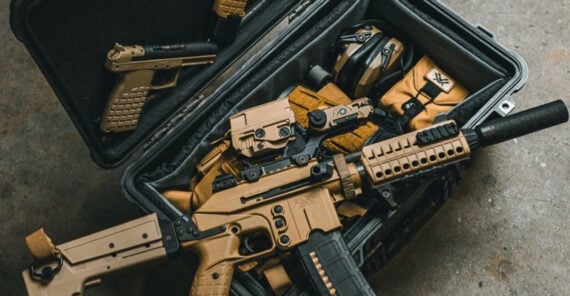 A black hard case on a concrete floor contains tan-colored firearms, including a pistol and a rifle with attachments, along with tactical gear and ear protection.
