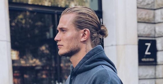 A man with blond hair tied in a low bun, wearing a blue hoodie, stands in profile against an urban backdrop with stone walls and a window.
