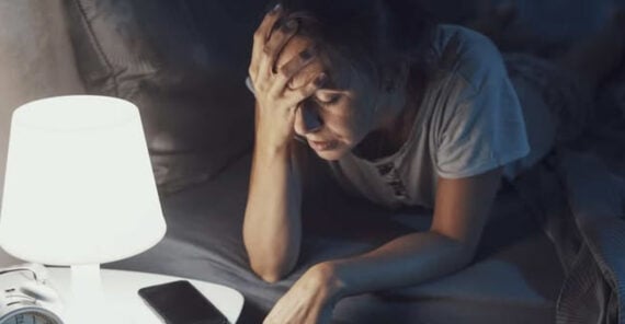 A woman lies in bed at night, looking stressed with her hand on her forehead. A lit lamp, a phone, and an alarm clock are on the bedside table. The room appears dim and she looks tired or worried.