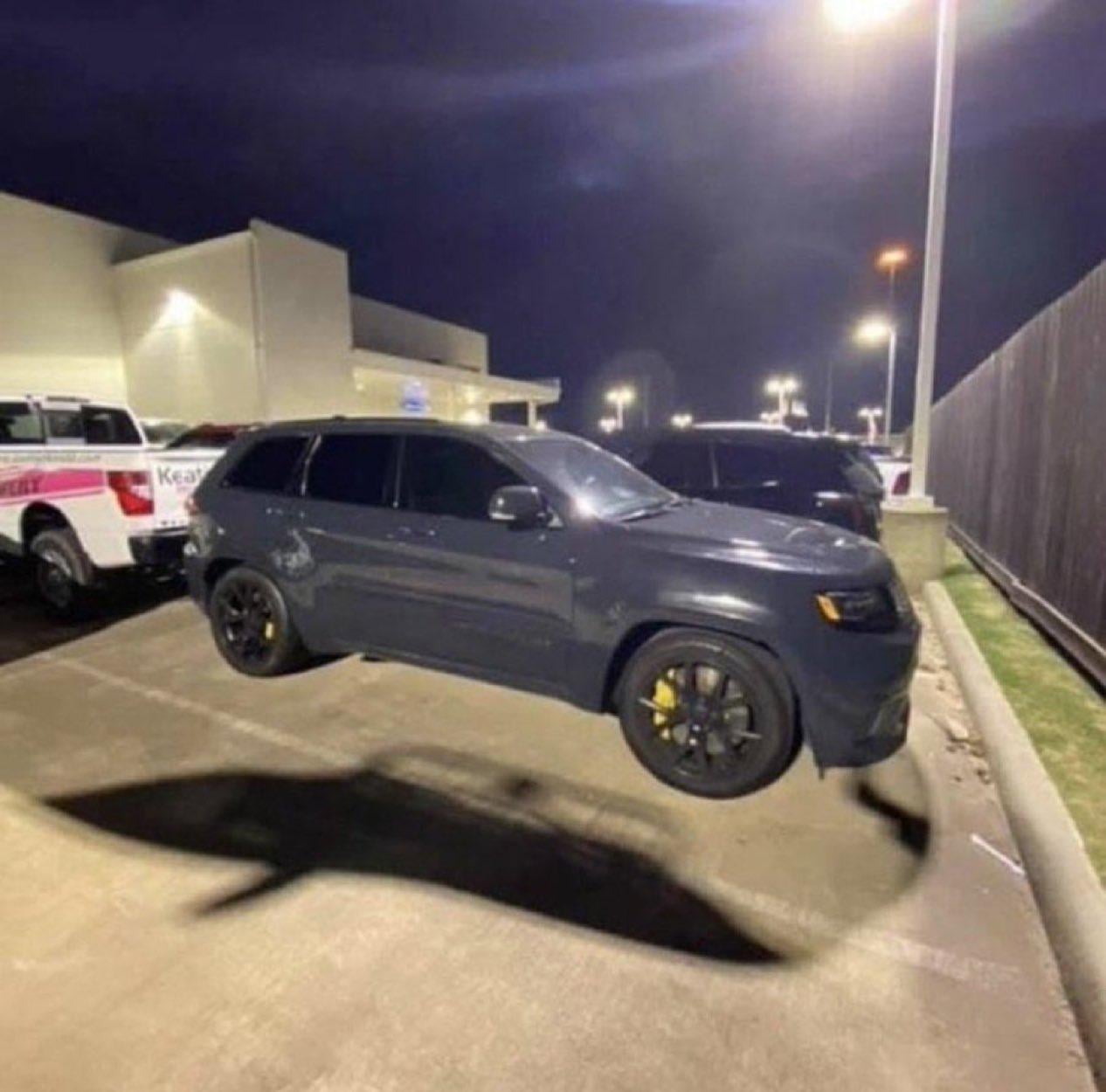 A black SUV with yellow brake calipers is parked in a lot at night. The streetlight casts a shadow beneath the car that resembles the shape of a shark. Other vehicles and a building are visible in the background.