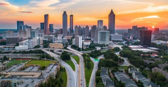 Aerial view of downtown Atlanta at sunset, showing skyscrapers, highways, green spaces, and residential buildings with a colorful sky in the background.