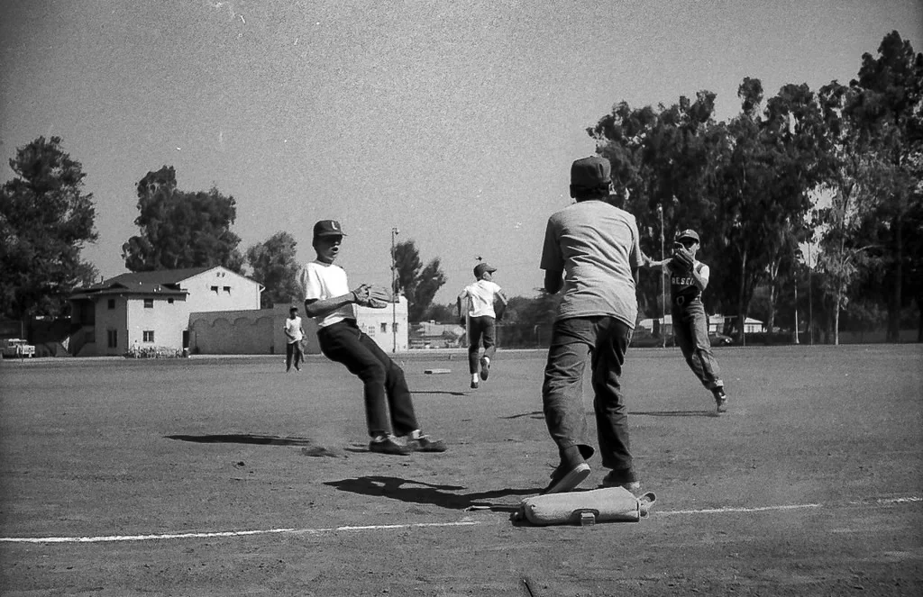 Black and white photo of boys playing baseball; one is sliding toward a base while another tries to tag him, with teammates and trees visible in the background on a sunny day.