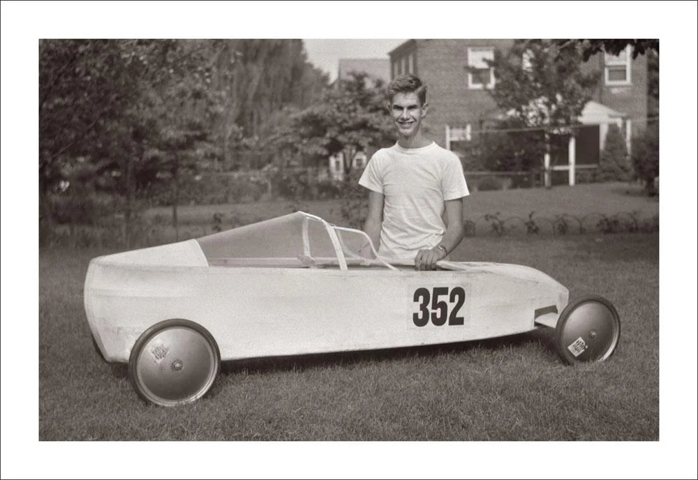 A young person in a white t-shirt stands beside a homemade, three-wheeled soapbox derby car numbered 352, outdoors on a grassy lawn with trees and a house in the background.