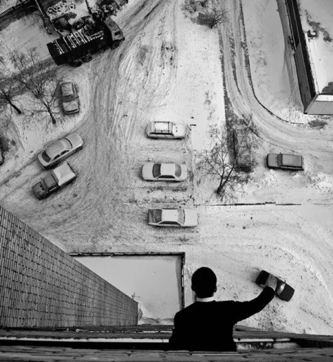 A person seen from behind leans out of a high window, waving over a snowy urban scene with several cars parked and tire tracks visible below.