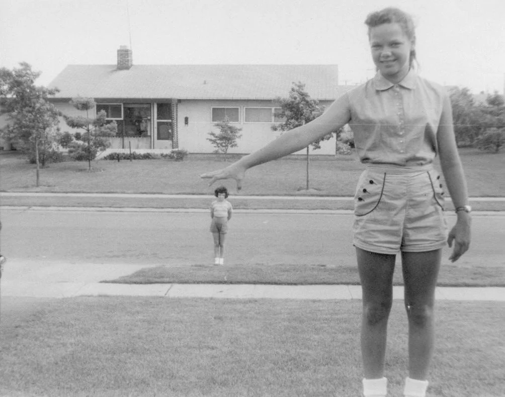 A black-and-white photo shows a girl in the foreground extending her arm toward a distant child across the street, creating an optical illusion that the girl is much larger than the child. A house and trees are in the background.