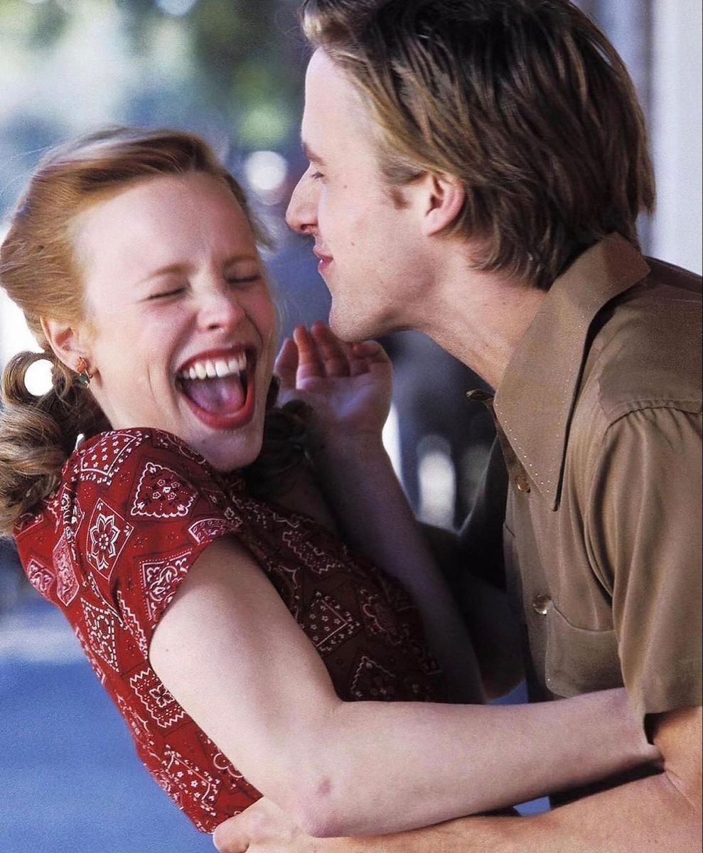 A woman in a red patterned dress laughs joyfully as a man in a brown shirt playfully holds her and leans in close, both appearing happy and carefree outdoors.